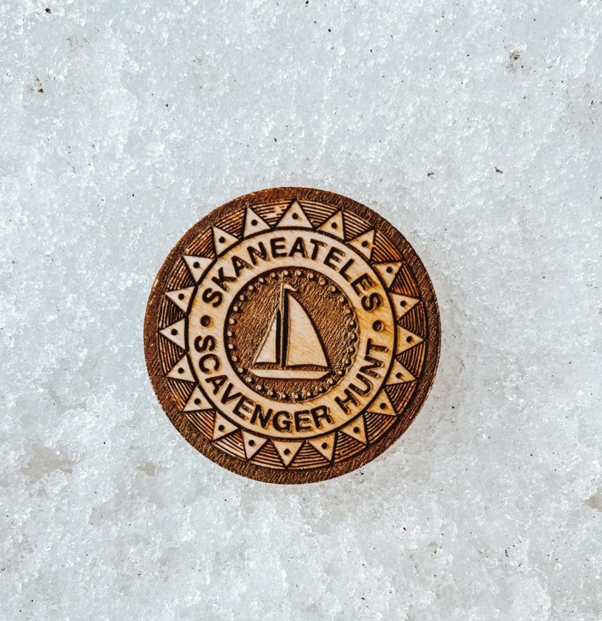 A wooden brown token on the snow - the token says Skaneateles Scavenger Hunt with a sailboat on it